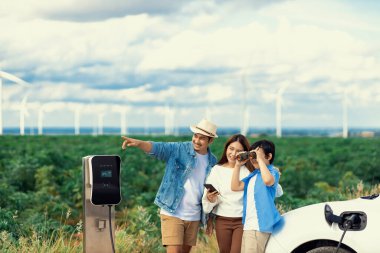 İlerici mutlu aile kavramı elektrikli araçla rüzgar çiftliğinde eğleniyor. Rüzgar türbini jeneratöründen gelen temiz yenilenebilir enerji ile şarj edilen elektrikli araç..