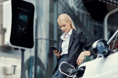 Businesswoman wearing black suit using smartphone, leaning on electric car recharge battery at charging station in city residential building with condos and apartment. Progressive lifestyle concept.