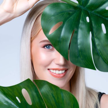 Closeup facial portrait personable woman with perfect smooth makeup holding green monstera leaves and cover her face as natural healthy skincare treatment. Tropical nature and beauty concept