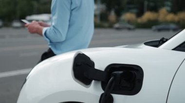 Takım elbiseli, hırslı bir işadamı elektrik kablosu ve yenilenebilir enerji enerjili bir elektrikli araçla şarj istasyonunda dururken elektrikli aracına yaslanıyor..