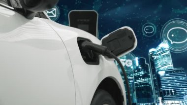 5G Network dijital hologramı ve internet tabelaları gece şehrinin arka planında beliriyor. Çevre koruma kavramı için kablo prizi olan şarj istasyonuna bağlı ilerici elektrikli araç.