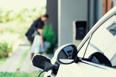 Focus EV car recharging at home charging station with blurred progressive woman and young girl in background for alternative clean energy technology concept for renewable electric vehicle.