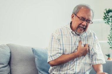 An agonizing senior man suffering from chest pain or heart attack alone in his living room. Serious health problem and feeling unwell concept for seniors.