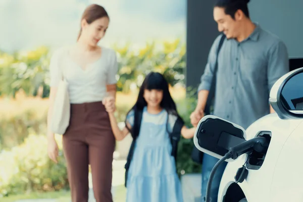 Stock image Focus closeup electric vehicle recharging battery from home electric charging station with blurred family in background. Renewable clean energy car for progressive eco awareness lifestyle concept.