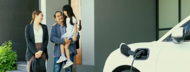 Progressive young parents and daughter with electric vehicle and home charging station. Green and clean energy from electric vehicles for healthy environment. Eco power from renewable source at home.