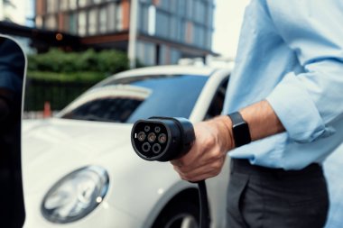 Closeup progressive man holding EV charger plug from public charging station for electric vehicle with background of residential building as concept eco-friendly sustainability energy car concept.