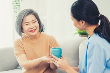 Female care taker serving her contented senior patient with a cup of coffee at home, smiling to each other. Medical care for pensioners, Home health care service.