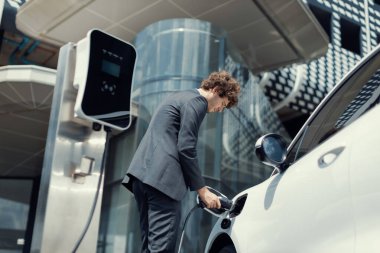 Below view closeup image of progressive black suit businessman recharge battery of his electric vehicle from public charging station. Renewable and alternative energy powered car concept.
