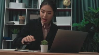 Kulaklık ve mikrofon takan iş kadını masasında çalışıyor ve dizüstü bilgisayar kullanıyor. Uzaktan kumandalı online toplantı için dizüstü bilgisayar kullanan hevesli kadın çalışanlar, ofiste iş videosu konferansı.
