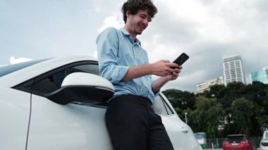 Takım elbiseli, hırslı bir işadamı elektrik kablosu ve yenilenebilir enerji enerjili bir elektrikli araçla şarj istasyonunda dururken elektrikli aracına yaslanıyor..