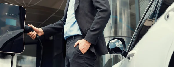 stock image Closeup progressive black suit businessman insert EV charger plug from public charging station to his electric car. Eco friendly electric rechargeable car with zero CO2 emission.