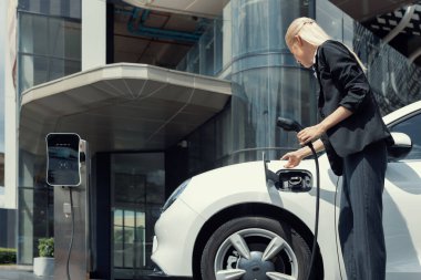 Progressive businesswoman insert charger plug from charging station to her electric vehicle with apartment condo building in background. Eco friendly rechargeable car powered by sustainable energy.