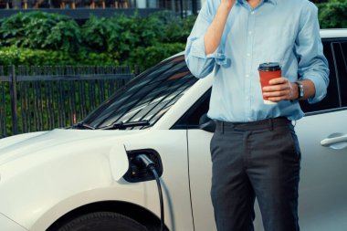 Progressive eco-friendly concept of parking EV car at public electric-powered charging station in city with blur background of businessman leaning on recharging-electric vehicle with coffee.