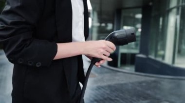 İlerici iş kadını şehir merkezinde dolaşmadan önce şarj cihazından elektrikli arabasına şarj prizi takıyor. Çevre dostu şarj edilebilir araba, sürdürülebilir ve temiz enerjiyle çalışıyor..