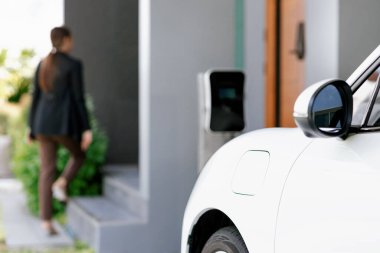 Focus image of electric vehicle recharging battery at home charging station with blurred woman walking in the background. Progressive concept of green energy technology applied in daily lifestyle.