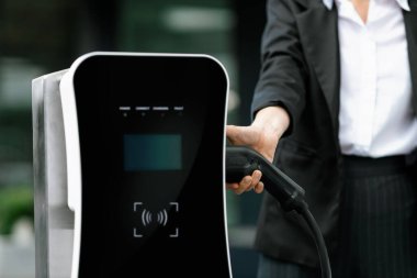 Focus EV charger plug and electric car at public charging station with blur progressive businesswoman holding charger and apartment condo building in background. Eco friendly electric vehicle concept.