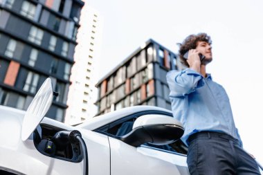 Progressive businessman talking on the phone, leaning on electric car recharging with public EV charging station, apartment condo residential building on the background as green city lifestyle.