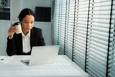 Competent female employee sits at her desk with a cup of coffee. Modern employee working with a drink, recreation during working hours, caffeine for people who are working