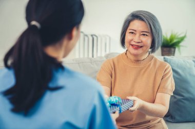 A young caregiver hand over to her senior patient a blue gift box with blue ribbons at a contented living room.