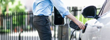 Closeup progressive man holding EV charger plug from public charging station for electric vehicle with background of residential building as concept eco-friendly sustainability energy car concept.