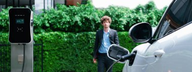 Progressive businessman wearing black suit with electric car recharging at public parking car charging station at modern city residential area. Eco friendly rechargeable car powered by clean energy.