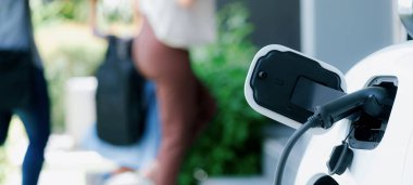 Focus closeup electric vehicle recharging battery from home electric charging station with blurred family in background. Renewable clean energy car for progressive eco awareness lifestyle concept.