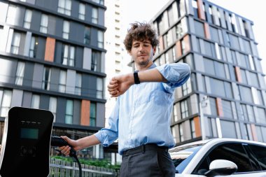 Businessman with smartwatch at modern charging station for electric vehicle with background of residential buildings as concept for progressive lifestyle of using eco-friendly as alternative energy.
