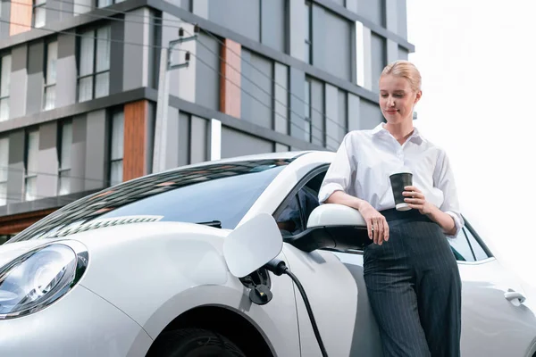 Businesswoman Drinking Coffee Leaning Electric Vehicle Recharging Public Charging Station — Stock fotografie