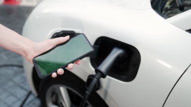 Taklit uzay için şarj istasyonuna bağlı elektrikli aracın yeşil ekran enerji durumunu gösteren bir model telefon. Temiz çevre için ilerici bir kavram. EV, yeşil yenilenebilir enerjiden güç alıyor.