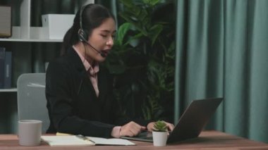 Kulaklık ve mikrofon takan iş kadını masasında çalışıyor ve dizüstü bilgisayar kullanıyor. Uzaktan kumandalı online toplantı için dizüstü bilgisayar kullanan hevesli kadın çalışanlar, ofiste iş videosu konferansı.