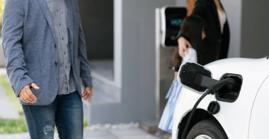 Closeup focus hand insert EV charger plug to electric vehicle at home charging station with blurred family in background. Progressive concept of green and clean energy to reduce CO2 emission by EV car