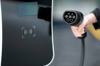 Focus closeup hand holding EV plug for electric vehicle pointing at camera with charging station at background as progressive idea of alternative sustainable clean energy for ecological awareness idea