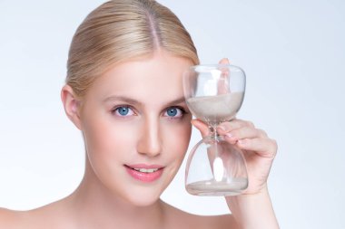 Closeup personable model holding hourglass in beauty concept of anti-aging skincare treatment. Young girl portrait with perfect smooth clean skin and flawless soft makeup in isolated background