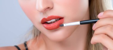 Closeup beautiful young woman with flawless healthy skin and natural makeup putting alluring fashion glossy red lipstick on her lip with lip brush in isolated background.