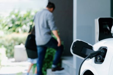 Arka planda bulanık baba ve kızının yürüdüğü şarj istasyonunda elektrikli araba şarj etmeye odaklan. Sağlıklı çevre yaşam tarzı konsepti için yenilikçi yeşil ve temiz enerji aracı.