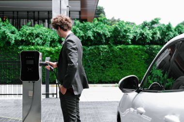 Modern şehir yerleşim bölgesindeki kamu şarj istasyonunda elektrikli araba şarj eden yenilikçi bir işadamı. Çevre dostu şarj edilebilir araba alternatif temiz enerjiyle çalışıyor..