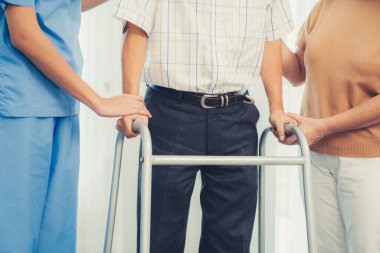 Contented senior man walking as he is helped by his wife and caretaker, walking with the aid of a folding walker. Nursing home for the elderly concept.