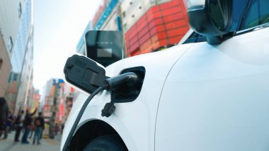 Şehir merkezindeki şarj istasyonundan enerji alan elektrikli araç, şehir merkezinde sokakta yürüyen insanlar. Temiz gelecek ortamı için sürdürülebilir enerji tarafından sürülen yenilikçi EV otomobil.