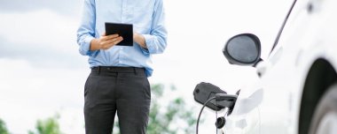 Closeup progressive suit-clad businessman with his electric vehicle recharge his car on public charging station in modern city with power cable plug and renewable energy-powered electric vehicle.