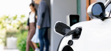 Focus closeup electric vehicle recharging battery from home electric charging station with blurred family in background. Renewable clean energy car for progressive eco awareness lifestyle concept.