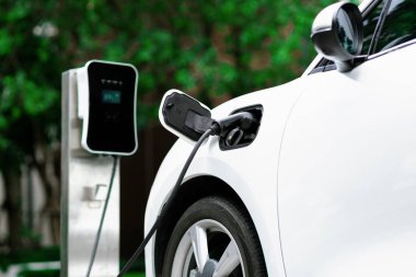 Focus closeup electric vehicle plugged in with EV charger device from blurred background of public charging station powered by renewable clean energy for progressive eco-friendly car concept.