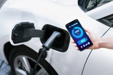 Battery status of electric vehicle displayed on smartphone application or software while vehicle is plugged into charging station for progressive future refueling. Battery status on phone screen.