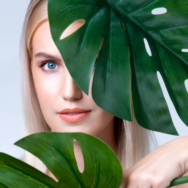 Closeup facial portrait personable woman with perfect smooth makeup holding green monstera leaves and cover her face as natural healthy skincare treatment. Tropical nature and beauty concept