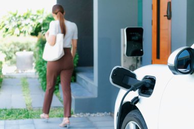 Focus EV charger plugged into EV car at home charging station with blurred background of progressive woman walking in background. Elective vehicle powered by clean energy for eco-friendly concept.