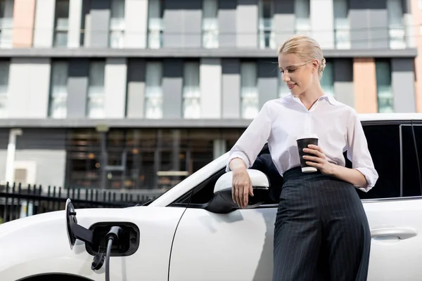 Businesswoman Drinking Coffee Leaning Electric Vehicle Recharging Public Charging Station — 图库照片