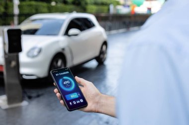 Closeup progressive businessman look at EV cars battery status application on smart phone screen at public parking charging station with power cable plug and renewable energy-powered electric vehicle