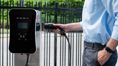 Closeup progressive man holding EV charger plug from public charging station for electric vehicle with background of residential building as concept eco-friendly sustainability energy car concept.