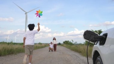 İlerici mutlu aile kavramı elektrikli araçla rüzgar çiftliğinde eğleniyor. Rüzgar türbini jeneratöründen gelen temiz yenilenebilir enerji ile şarj edilen elektrikli araç..