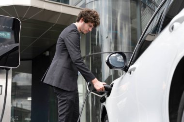 Below view closeup image of progressive black suit businessman recharge battery of his electric vehicle from public charging station. Renewable and alternative energy powered car concept.