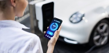Suit-clad progressive businesswoman look at EV cars battery status from her phone while standing on a charging station with a power cable plug and a renewable energy-powered electric vehicle.